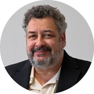 Ein Mann mit grauen Haaren und einem Bart lächelt freundlich in die Kamera. Er trägt einen schwarzen Blazer und ein helles Oberteil. Der Hintergrund ist neutral und hell. Die Aufnahme ist in einem runden Format präsentiert.