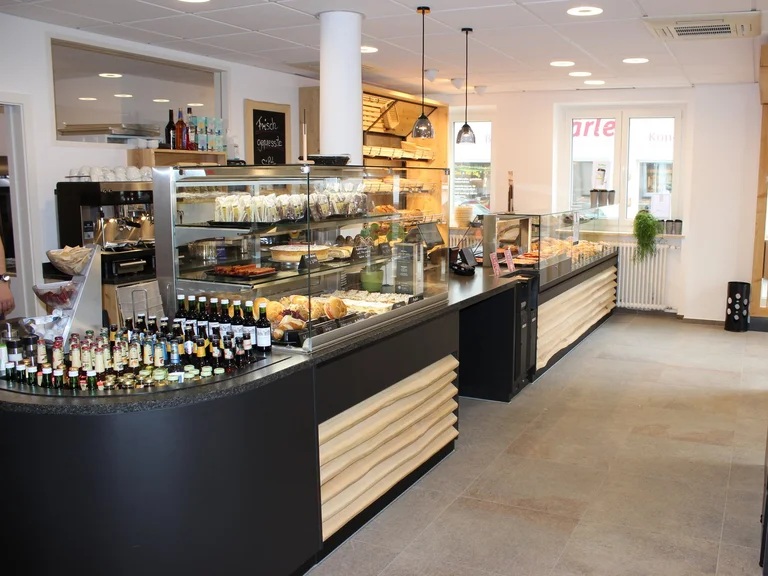 Eine moderne Cafeteria mit einer Theke. Vor der Theke sind verschiedene Gebäckstücke, Snacks und Getränke in Glasvitrinen präsentiert. Die Umgebung ist hell und freundlich gestaltet, mit Holzelementen und einer klaren, strukturierten Anordnung.