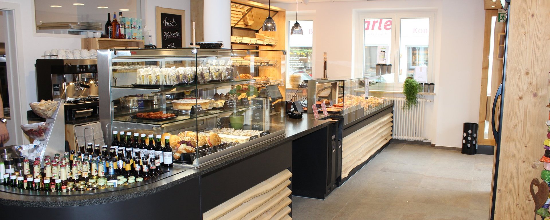 Eine moderne Cafeteria mit einer Theke. Vor der Theke sind verschiedene Gebäckstücke, Snacks und Getränke in Glasvitrinen präsentiert. Die Umgebung ist hell und freundlich gestaltet, mit Holzelementen und einer klaren, strukturierten Anordnung.