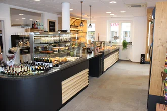 Eine moderne Cafeteria mit einer Theke. Vor der Theke sind verschiedene Gebäckstücke, Snacks und Getränke in Glasvitrinen präsentiert. Die Umgebung ist hell und freundlich gestaltet, mit Holzelementen und einer klaren, strukturierten Anordnung.