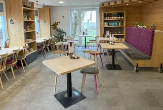 Ein gemütlicher Innenraum eines Cafés mit hellen Holzmöbeln, Tischen und Stühlen. Zu sehen sind einige Pflanzen sowie eine Vitrine mit dekorativen Gegenständen. Große Fenster lassen Tageslicht herein und bieten einen Blick nach draußen. Ein gepolsterter Sitzbereich ist ebenfalls vorhanden.