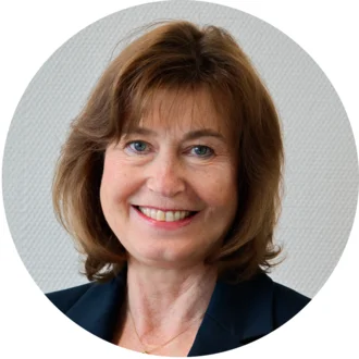 Ein Porträt einer Frau mit schulterlangem braunem Haar und blauer Jacke, die lächelt. Ihr Gesicht ist freundlich und einladend, mit blauen Augen und einem leichten Schmuckstück am Hals. Der Hintergrund ist hell und neutral.