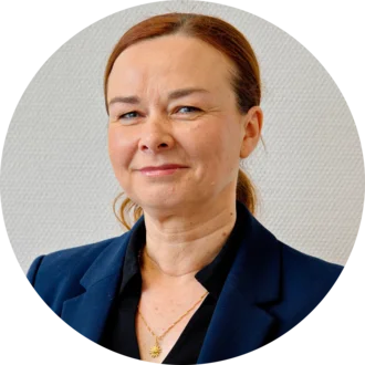 Porträt einer Frau mit schulterlangen, braunen Haaren, die in einem Dunkelblauen Blazer gekleidet ist. Sie schaut direkt in die Kamera und hat einen freundlichen Gesichtsausdruck. Der Hintergrund ist einheitlich und hell.