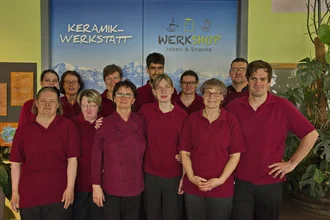 Eine Gruppe von zwölf Personen steht lächelnd vor einem Hintergrund mit den Schriftzügen "Keramik-Werkstatt" und "Werkshop Ideen & Snacks". Alle tragen rote Oberteile. Im Vordergrund sind drei Personen in einem engen Halbkreis angeordnet, während die anderen im Hintergrund stehen.