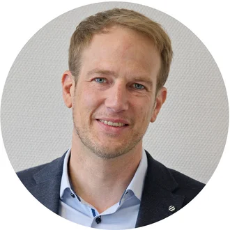 Porträt eines lächelnden Mannes mit kurzen, hellblonden Haaren und einem leichten Bart. Er trägt ein blaues Jackett über einem hellblauen Hemd. Der Hintergrund ist neutral und strukturiert.
