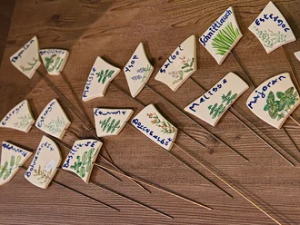 Eine Reihe von kleinen, dreieckigen Schildern aus Keramik auf Holzuntergrund, die beschriftet sind mit Namen von Kräutern wie "Dill", "Majoran", "Schnittlauch" und "Melisse". Jedes Schild hat eine Pflanzenillustration und ist auf einem langen Stab befestigt.