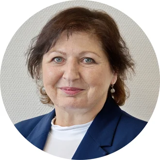 Porträt einer Frau mit kurzen, braunen Haaren und hellen Augen. Sie trägt eine blaue Jacke und einen weißen Oberteil. Der Hintergrund ist neutral und unscharf, um den Fokus auf ihr Gesicht zu legen.