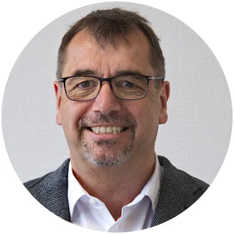 Porträt eines lächelnden Mannes mit Brille und Halbglatze, der einen grauen Blazer und ein weißes Hemd trägt. Der Hintergrund ist hell und strukturiert.