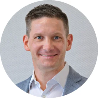Ein Mann mit kurzen, grauen Haaren und blauen Augen lächelt freundlich. Er trägt ein helles Hemd und eine graue Jacke. Der Hintergrund ist neutral und strukturiert, wodurch der Mann im Vordergrund hervorgehoben wird.