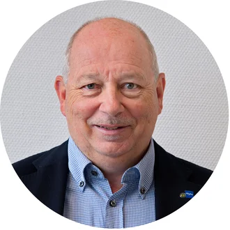 Ein älterer Mann mit einer leicht grauen Frisur lächelt freundlich. Er trägt ein blaues Hemd mit Karomuster und einen dunklen Blazer. Der Hintergrund ist hell und strukturiert. Der Mann wirkt sympathisch und offen.