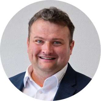 Ein Mann mit kurzen, lockigen Haaren und einem freundlichen Lächeln schaut in die Kamera. Er trägt ein Hemd mit Kragen und einen blauen Blazer. Der Hintergrund ist hell und neutral.