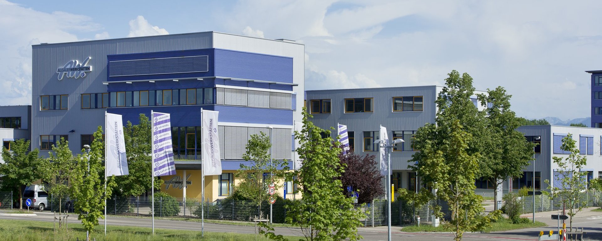 Das Bild zeigt ein modernes, mehrstöckiges Gebäudekomplex mit blauem und grauem Design. Vor dem Gebäude stehen mehrere Fahnen und es sind Bäume in der Umgebung sichtbar. Der Himmel ist klar mit einigen Wolken.
