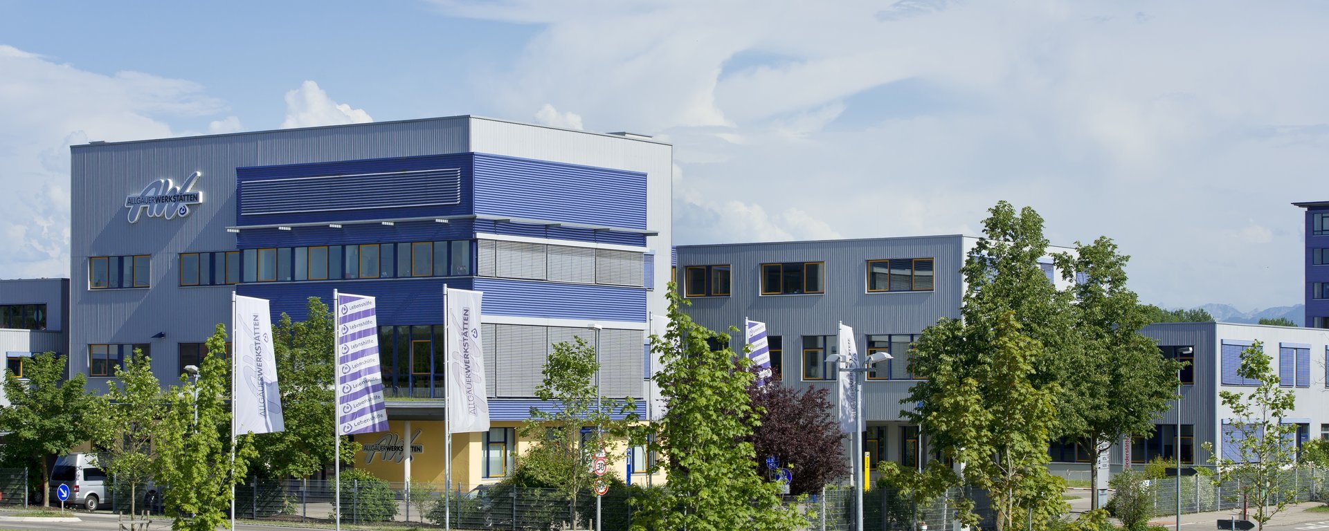 Das Bild zeigt ein modernes, mehrstöckiges Gebäudekomplex mit blauem und grauem Design. Vor dem Gebäude stehen mehrere Fahnen und es sind Bäume in der Umgebung sichtbar. Der Himmel ist klar mit einigen Wolken.