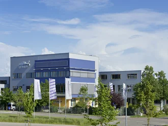 Das Bild zeigt ein modernes, mehrstöckiges Gebäudekomplex mit blauem und grauem Design. Vor dem Gebäude stehen mehrere Fahnen und es sind Bäume in der Umgebung sichtbar. Der Himmel ist klar mit einigen Wolken.