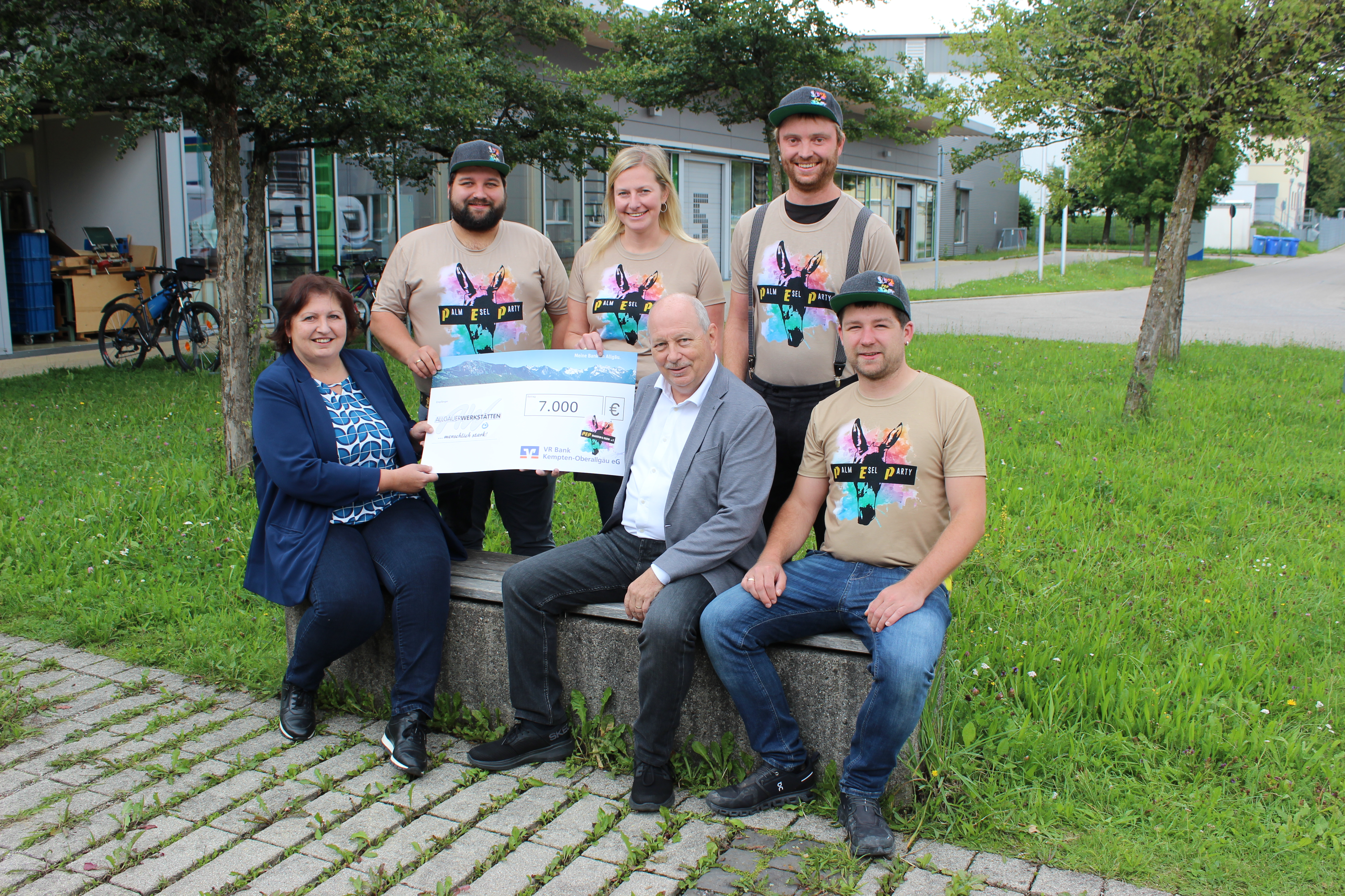 Eine Frau und fünf Männer stehen und sitzen gemeinsam auf einer Bank. Sie halten einen Scheck über 7.000 Euro, der an eine Organisation gerichtet ist. Alle tragen T-Shirts mit buntem Design. Im Hintergrund ist viel grüne Wiese und ein Gebäude sichtbar.
