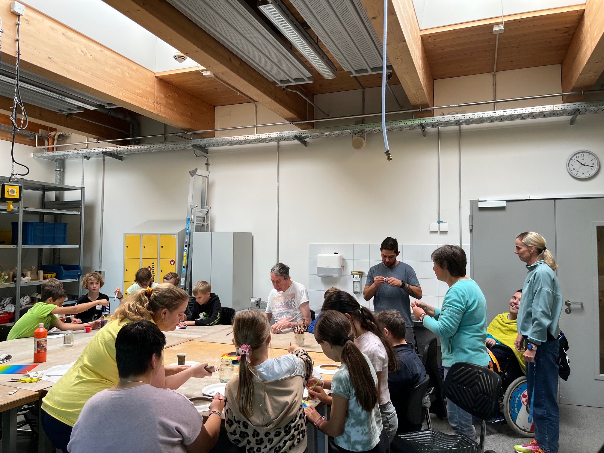 In einem hellen Raum arbeiten Kinder und Erwachsene an einem Tisch, um verschiedene kreative Projekte zu gestalten. Einige Personen sitzen, andere stehen, während sie Materialien wie Farbe und Pinsel nutzen. Es herrscht eine freundliche und aktive Atmosphäre.