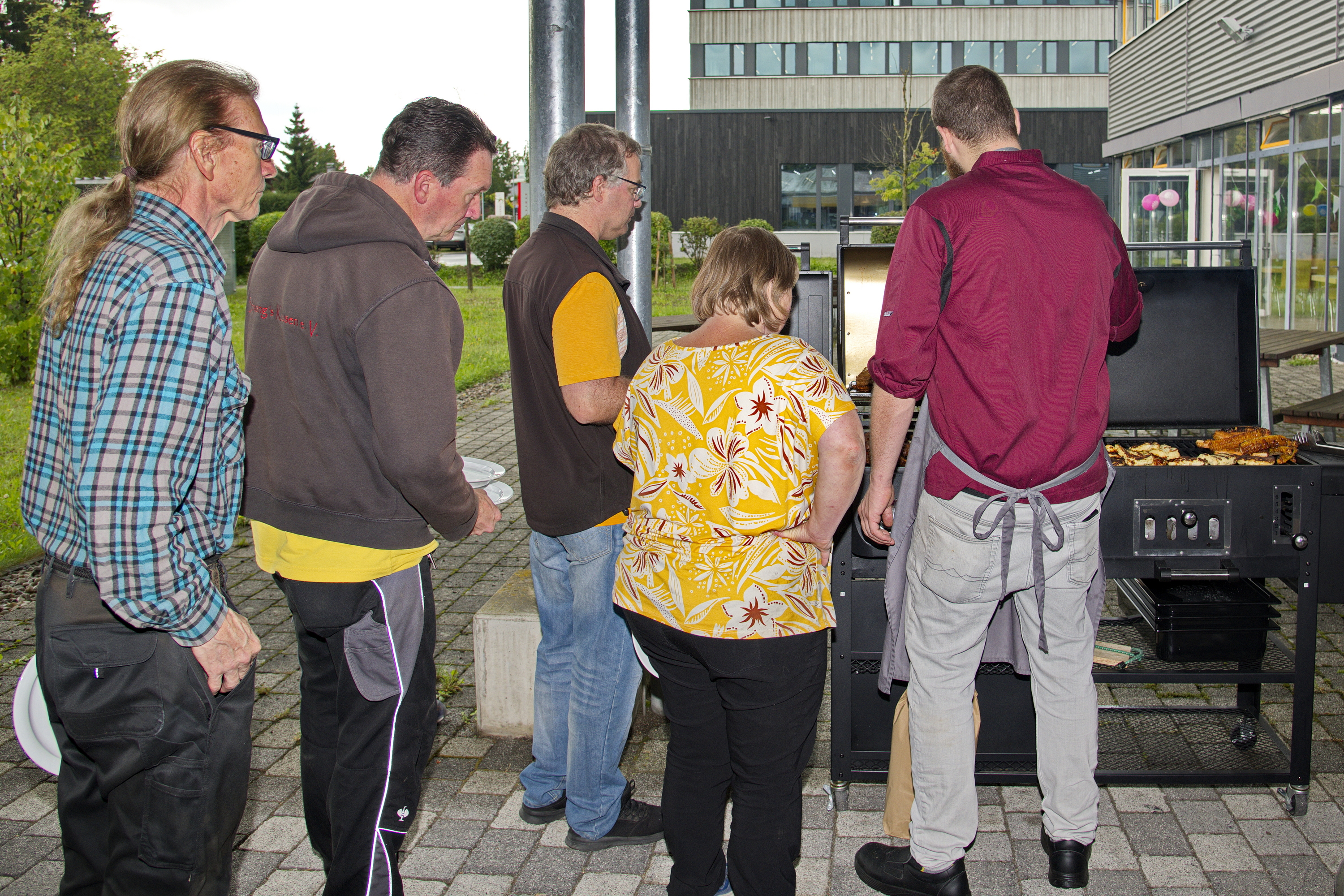 Eine Gruppe von fünf Personen steht an einem Grill im Freien. Ein Mann in einem roten Hemd grillt, während die anderen aufmerksam zuschauen. Sie scheinen an einem geselligen Grillabend teilzunehmen. Im Hintergrund sind Bäume und ein Gebäude zu sehen.